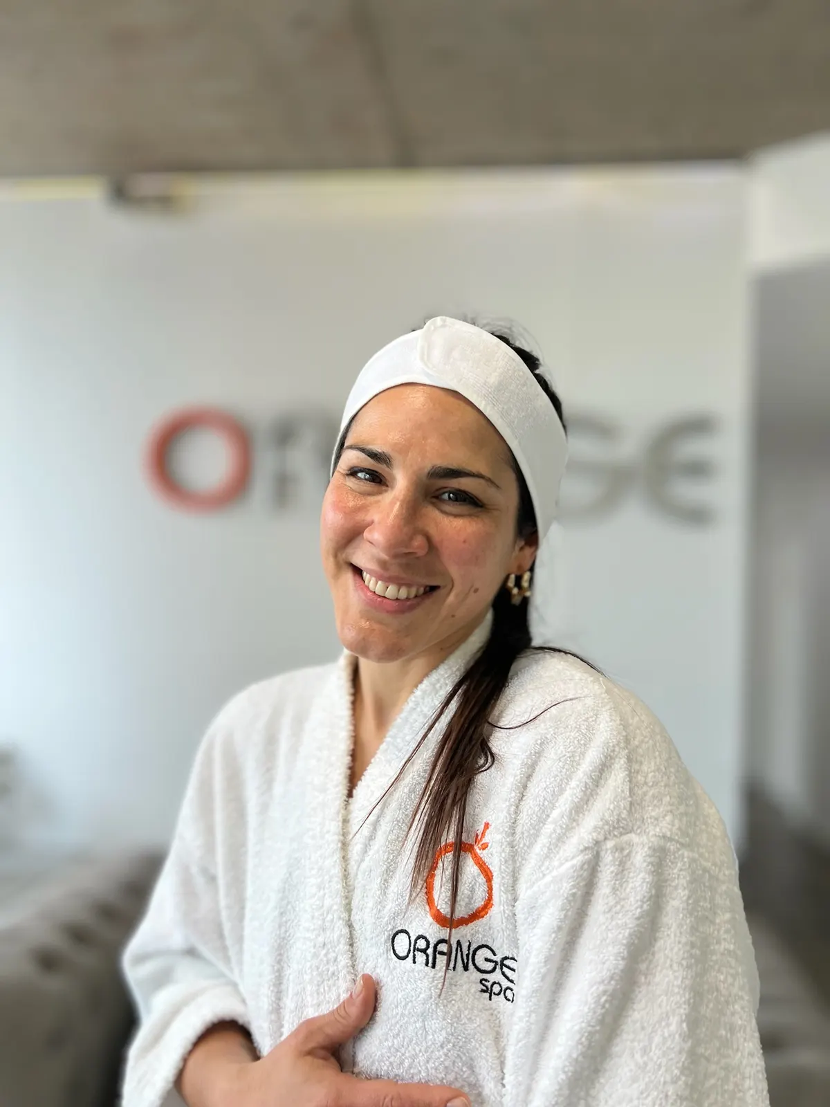 Clienta sonriente con bata de Orange Spa y vincha frente al cartel del centro