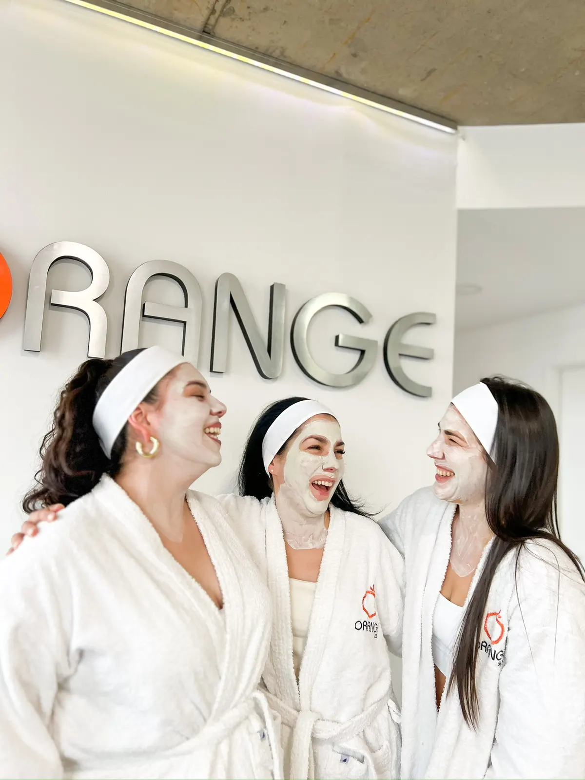 Tres clientas con batas y máscaras faciales riéndose frente al cartel de Orange